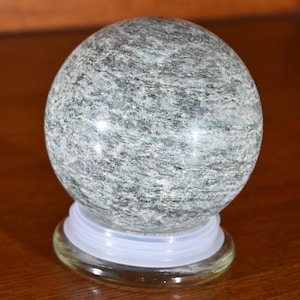 May include: A smooth, green and gray marble sphere on a clear glass base. The sphere has a swirling pattern.