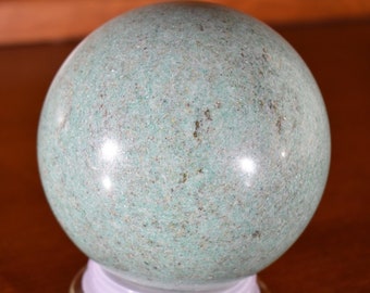 May include: A smooth, green sphere made of polished stone. The sphere is sitting on a clear glass base.