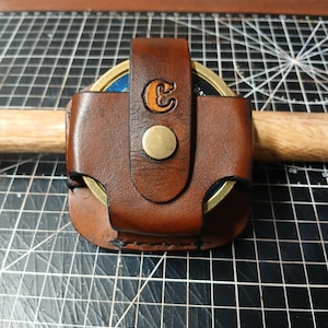 Könnte beinhalten: Ein handgefertigter brauner Leder-Münzhalter mit einem Messingknopf und einem gestempelten "C" auf der Klappe. Der Halter ist so konzipiert, dass er eine runde Münze umschließt und auf einem Holzdübel ruht.