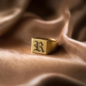 Könnte beinhalten: Ein goldfarbener Siegelring mit einer flachen, rechteckigen Oberfläche, in die ein dunkler Buchstabe R in altenglischer Schrift eingraviert ist. Der Ring hat eine gebürstete Metalloberfläche und eine massive Ringschiene mit einem klaren Design.