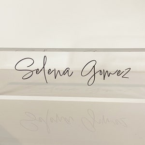 May include: A clear acrylic block with the name "Selena Gomez" written in black cursive script.