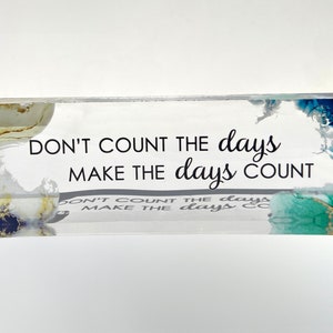 May include: A clear acrylic block with a blue, green, and gold abstract design. The block has the quote "Don't count the days, make the days count" printed on it.