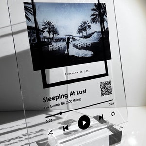 Puede incluir: Un marco de fotos de acrílico transparente con un borde negro, que presenta una foto en blanco y negro de una pareja de recién casados. El marco tiene un diseño de reproductor de música con el texto "Sleeping At Last" y "I'm Gonna Be (500 Miles)" junto con un botón de reproducción, un botón de retroceso y un código QR.