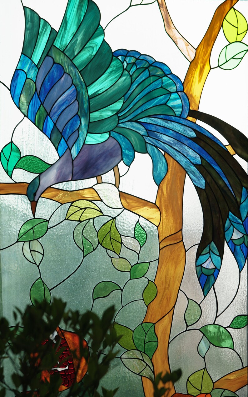 May include: Stained glass artwork featuring a bird with teal, blue, and purple plumage perched on a tree branch. The bird is surrounded by green leaves and a brown tree trunk, with a white background.