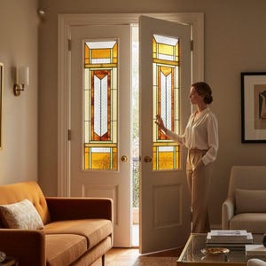 Puede incluir: Interior de un salón con una mujer cerca de puertas dobles. Las puertas presentan vidrieras con diseños geométricos en tonos amarillos, naranjas y blancos. Un sofá naranja y sillas de tonos neutros completan la decoración, creando un ambiente cálido.