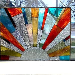 May include: Stained glass panel featuring a sunburst design. The artwork incorporates various colors, including red, orange, yellow, blue, and silver, arranged in a geometric pattern. The sun's rays emanate from a semi-circular base, creating a vibrant and decorative piece.