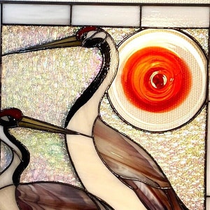 May include: A stained glass window depicting two cranes with a red and orange sun in the background. The cranes are made of iridescent glass and have black and white markings.