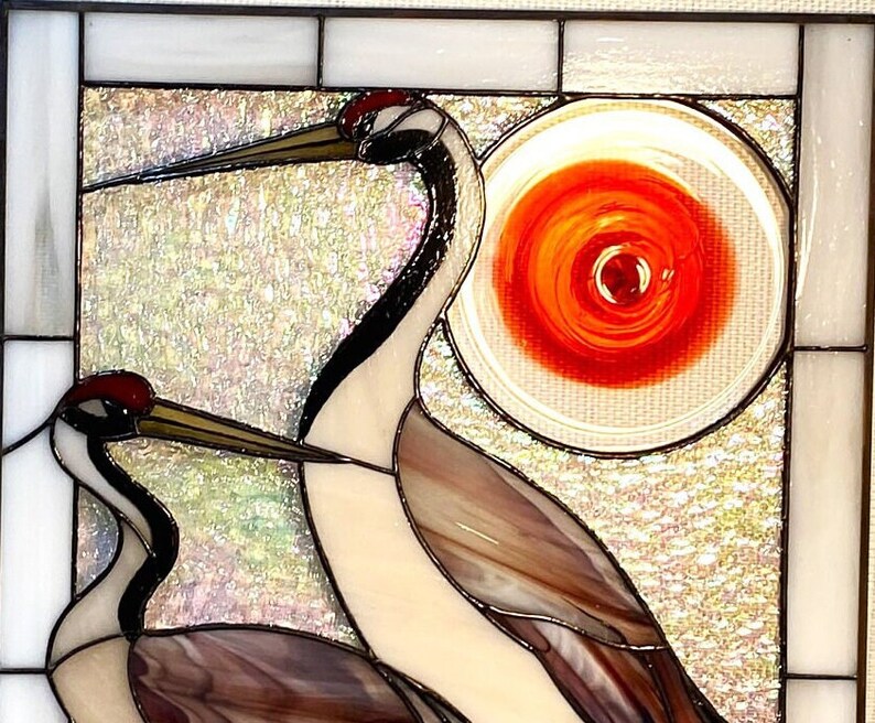 May include: A stained glass window depicting two cranes with a red and orange sun in the background. The cranes are made of iridescent glass and have black and white markings.