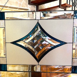 May include: Stained glass window with a geometric design. The window features a central diamond shape with teal and clear glass, surrounded by rectangular and square panels. The frame is black, and the glass has a textured appearance.