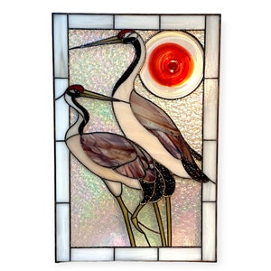 May include: A stained glass window panel depicting two cranes standing in front of a red sun. The cranes are white with black markings and long legs. The background is a shimmering iridescent white.