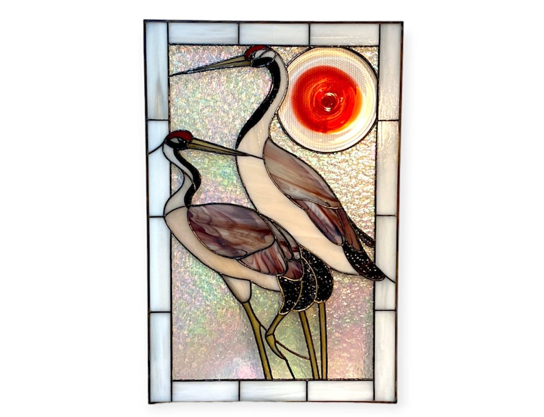May include: A stained glass window panel depicting two cranes standing in front of a red sun. The cranes are white with black markings and long legs. The background is a shimmering iridescent white.
