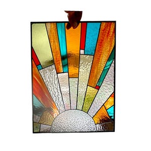 May include: A stained glass window panel depicting a stylized sun with a geometric design. The sun is made up of various colored glass pieces, including yellow, orange, red, blue, and green. The background is a clear glass with a textured surface.