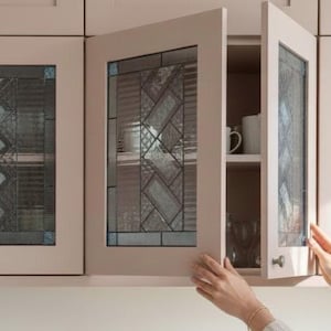 Op de afbeelding: Crèmekleurige keukenkasten met glazen deuren. Het glas heeft een decoratief loodontwerp in grijstinten en blauw. Eén kastdeur is open en onthult planken met witte mokken en glaswerk.