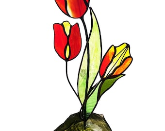 May include: A stained glass sculpture of three tulips in shades of red, orange, and yellow. The flower stems and leaves are green. The tulips are mounted on a textured, gold-coloured base. The artwork is set against a white background.