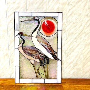 May include: Stained glass window panel featuring two cranes with long necks and legs, standing in front of a red sun. The cranes are made of iridescent glass with brown, white, and black accents.