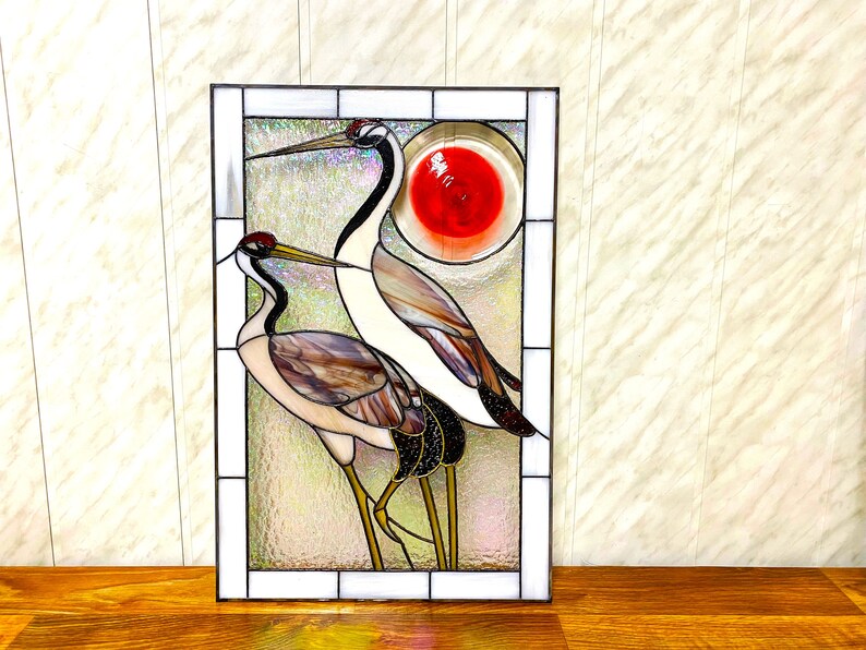 May include: Stained glass window panel featuring two cranes with long necks and legs, standing in front of a red sun. The cranes are made of iridescent glass with brown, white, and black accents.