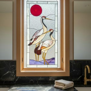 Puede incluir: Vidriera con dos grullas y un sol rojo. Las aves tienen plumas blancas, marrones y naranjas, de pie sobre una base morada y azul. La ventana está enmarcada en madera clara.