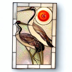 May include: A stained glass window panel depicting two cranes with a red sun in the background. The cranes are white with brown and black markings. The sun is a circle with a red swirl pattern.