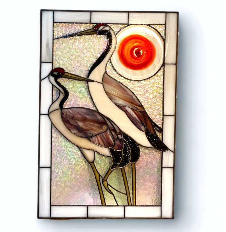 May include: A stained glass window panel depicting two cranes with a red sun in the background. The cranes are white with brown and black markings. The sun is a circle with a red swirl pattern.