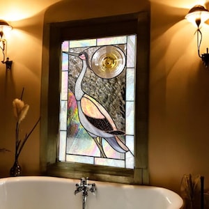 Puede incluir: Vidriera con diseño de grulla y elemento circular solar o lunar. El ave se representa en blanco, gris, rosa y negro. La ventana está enmarcada en madera, con un fondo texturizado. Ideal para decoración de baños.
