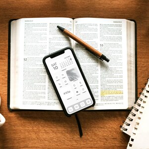 May include: A white ceramic mug, a brown leather-bound book, a black smartphone with a calendar app open, a brown pen, a white spiral notebook, and a pair of glasses on a wooden table.