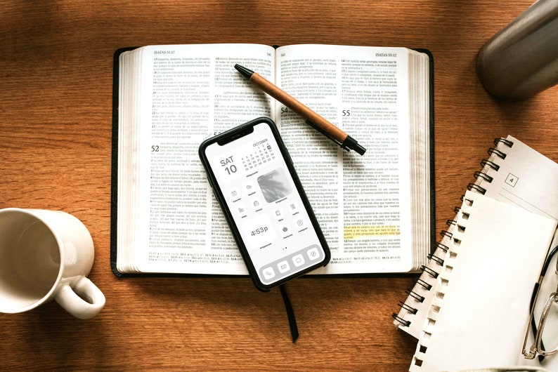 May include: A white ceramic mug, a brown leather-bound book, a black smartphone with a calendar app open, a brown pen, a white spiral notebook, and a pair of glasses on a wooden table.