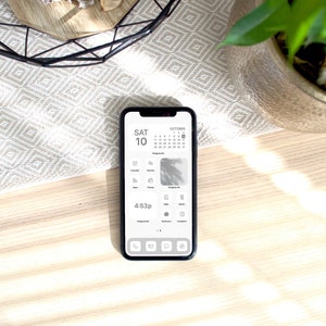 May include: A white iPhone with a black case displaying a calendar app with the date "SAT 10" and the time "4:63p". The phone is on a light wood surface with a white and gray patterned rug in the background.