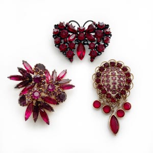 May include: Three vintage brooches with red and purple rhinestones. One is butterfly-shaped, another is a floral design, and the third is a round brooch with dangling teardrop-shaped gems. All are set on gold or black metal.