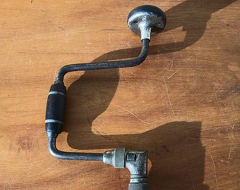 Op de afbeelding: Een vintage handboor met een zwarte houten handgreep en een metalen kruk. De boor heeft een metalen boorkop en een metalen tandwielmechanisme.