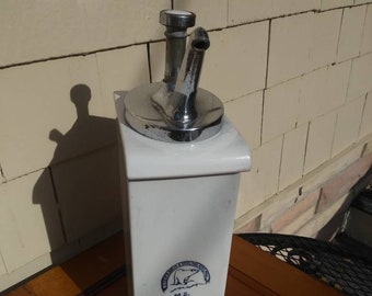 May include: A white ceramic dispenser with a chrome spout and top. The dispenser has a blue emblem with a polar bear and the initials "M.S." on the front. The dispenser is rectangular and sits on a wooden surface.