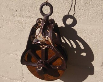 May include: A vintage, rusty, cast iron pulley with a wooden wheel. The pulley is hanging from a wire and has a loop at the top.