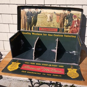 May include: Vintage Bruner Woolens display case. The dark green case features a colorful illustration of men in suits and the text "Quality Woolens for fine Custom Tailoring." The case also has the text "100% Pure Wool Stamp on Every Yard."