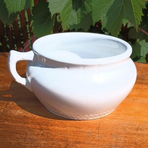 May include: A white ceramic bowl with a handle. The bowl has a rounded shape and a wide opening. The handle is curved and attached to the side of the bowl. The bowl is sitting on a wooden surface.