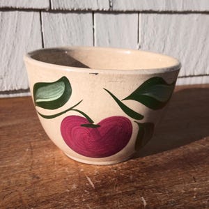 May include: Cream-colored ceramic bowl with a hand-painted design of a large, deep red cherry and green leaves. The bowl has a slightly flared rim and sits on a wooden surface. The cherry design is centered on the bowl's exterior.