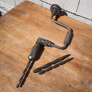 May include: An antique hand drill with a metal frame and wooden handle. Two drill bits are next to the drill. The drill and bits are made of metal and have a worn appearance. The drill is on a wooden surface.