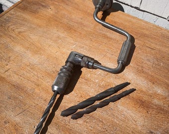May include: An antique hand drill with a metal frame and wooden handle. Two drill bits are next to the drill. The drill and bits are made of metal and have a worn appearance. The drill is on a wooden surface.