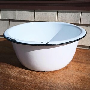 May include: A white enamelware bowl with a black rim. The bowl is round and has a slightly distressed appearance, suggesting it is vintage. The bowl sits on a wooden surface.