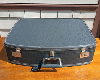 May include: A vintage black suitcase with a textured fabric exterior and a metal handle. The suitcase has two metal clasps on the sides and a metal latch on the front.