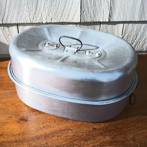 May include: A vintage, oval-shaped aluminum mess kit with a lid and handle. The kit is silver with a slightly worn appearance, suggesting age and use. The lid has two small latches and a black handle for easy carrying. The kit is sitting on a wooden surface.