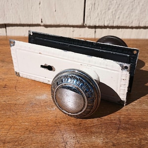 May include: Antique door hardware set featuring a silver-toned, ornate doorknob and a white and black door plate. The doorknob has a detailed, circular design. The door plate has a keyhole and decorative edges. The set is displayed on a wooden surface.
