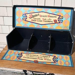 May include: Vintage display case with the text "Bruner Quality Woolens" and "Exclusive Fabrics." The case is black with a colorful interior featuring geometric patterns and stylized figures. The case is open, revealing three compartments.