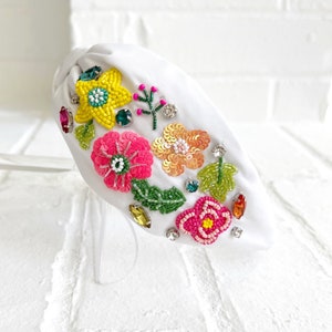 May include: White headband with a floral design. The headband is decorated with colorful beads, sequins, and rhinestones. The flowers are pink, yellow, and green.
