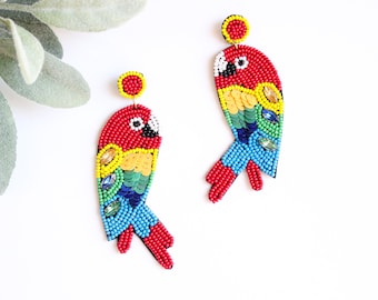 May include: A pair of beaded parrot earrings. The earrings are red, yellow, blue, and green, with black and white accents. The parrot earrings are on a white background with a green leafy plant in the upper left corner.