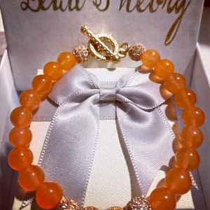 Puede incluir: Pulsera de piedras preciosas de color naranja con detalles dorados y cierre de palanca. La pulsera se muestra en una caja blanca con una cinta de sat&eacute;n blanca.