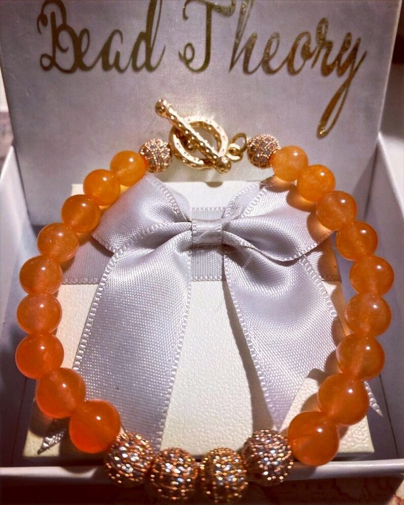 Puede incluir: Pulsera de piedras preciosas de color naranja con detalles dorados y cierre de palanca. La pulsera se muestra en una caja blanca con una cinta de sat&eacute;n blanca.
