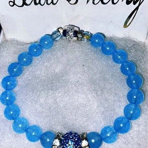 Puede incluir: Un brazalete de cuentas azul claro con un cierre plateado y un dije de cristal azul. El brazalete est&aacute; en una caja blanca con las palabras "Bead Theory" escritas en la parte superior.
