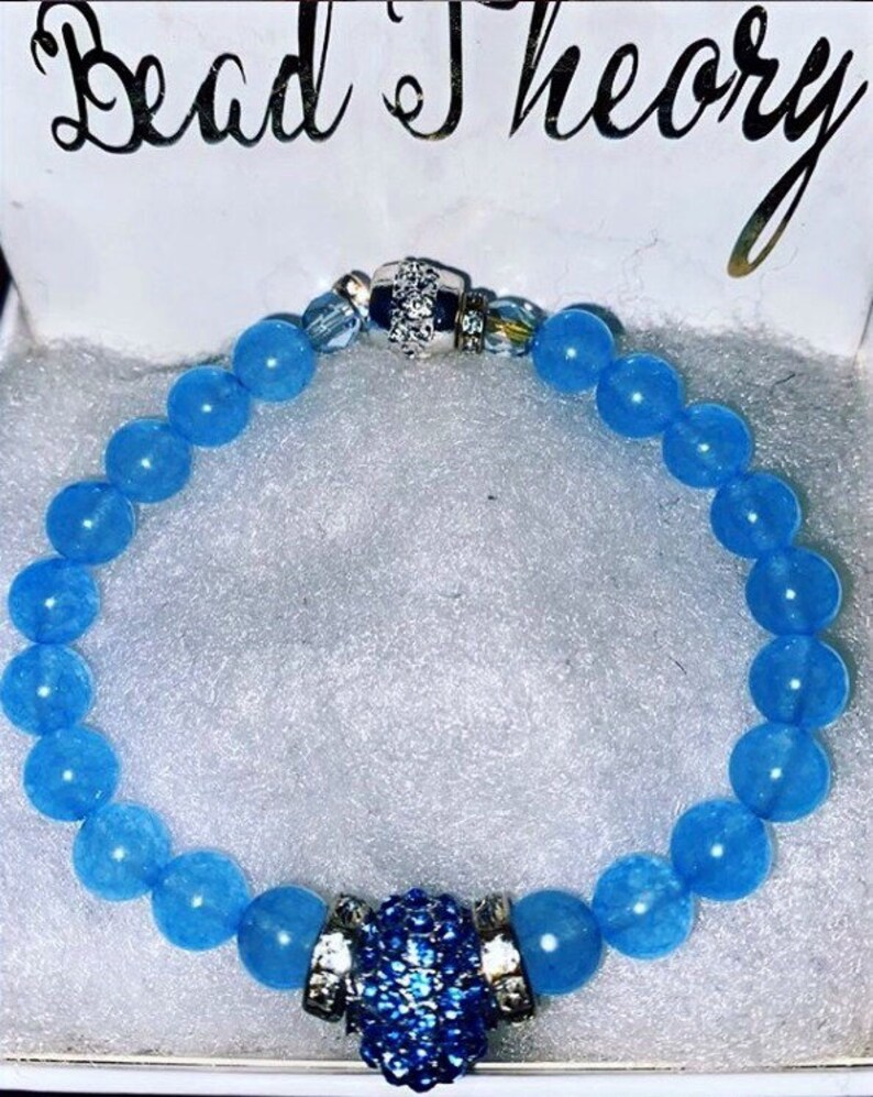 Puede incluir: Un brazalete de cuentas azul claro con un cierre plateado y un dije de cristal azul. El brazalete est&aacute; en una caja blanca con las palabras "Bead Theory" escritas en la parte superior.