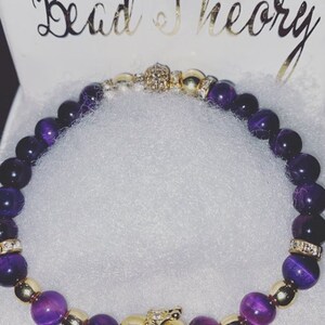 Puede incluir: Pulsera de cuentas moradas y doradas con un dije de corona dorada. La pulsera est&aacute; hecha con cuentas de ojo de tigre y separadores dorados.