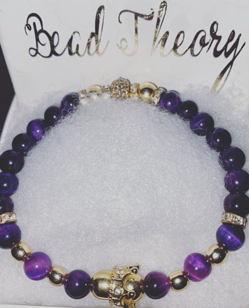 Puede incluir: Pulsera de cuentas moradas y doradas con un dije de corona dorada. La pulsera est&aacute; hecha con cuentas de ojo de tigre y separadores dorados.