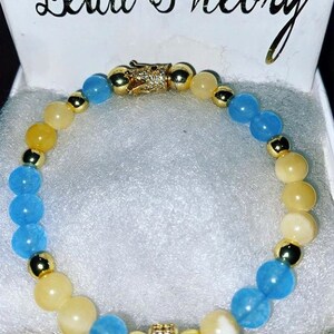 Puede incluir: Un brazalete de cuentas azules y amarillas con detalles dorados y un dije de corona. El brazalete est&aacute; en una caja blanca con las palabras "Bead Theory" impresas en la parte superior.